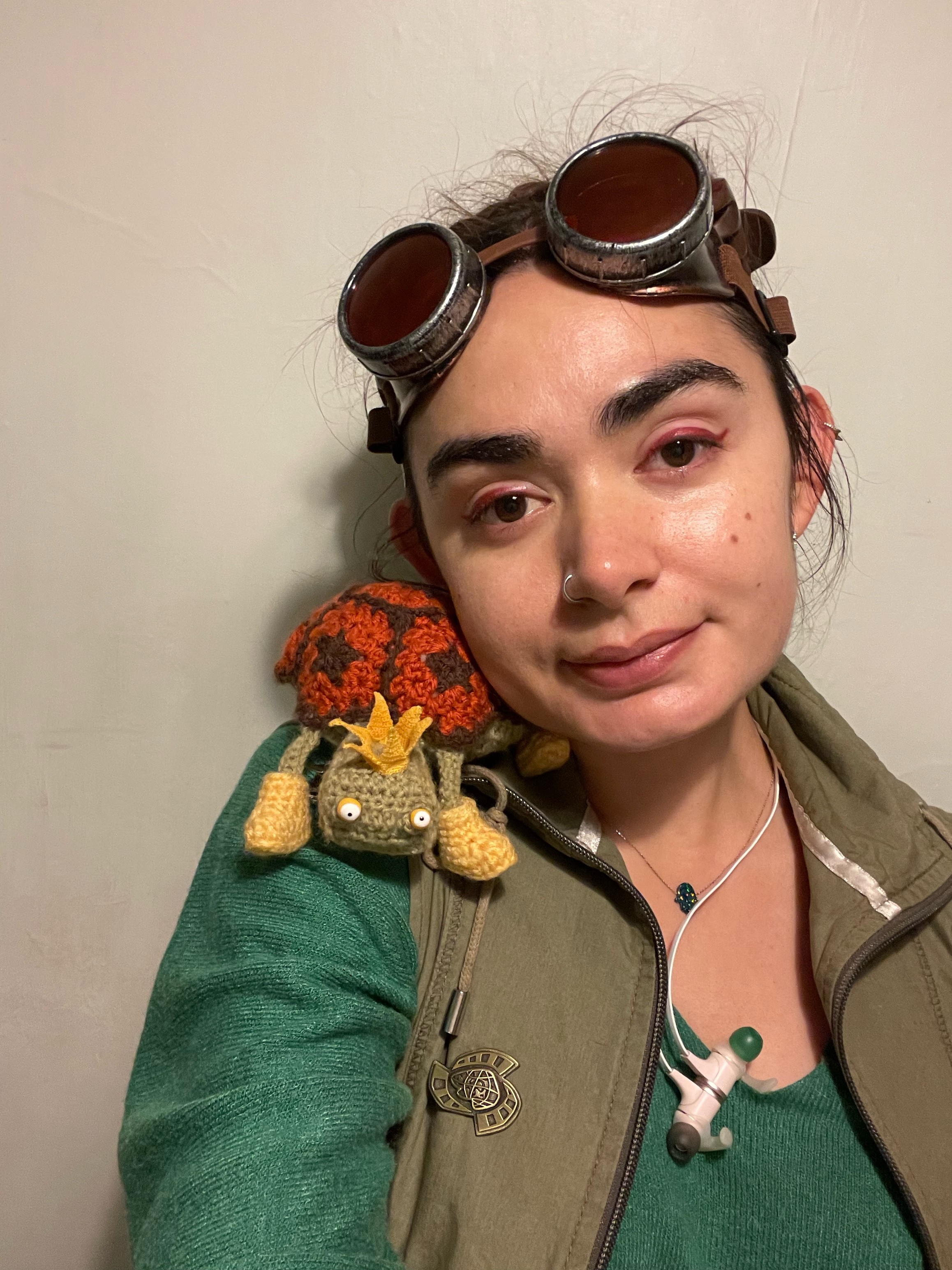 Leaning my head on mr pokeylope the tortoise plush like a pillow who is on my shoulder. I made him one eye bigger than the other and I printed the pupils slightly different too, he is also wearing a micro crocheted yellow crown and little yellow booties on the ends of his dangly little legs. His shell is orange and brown. I’m wearing my red-lensed goggles and my hair is a little wild with pieces sticking up at the top