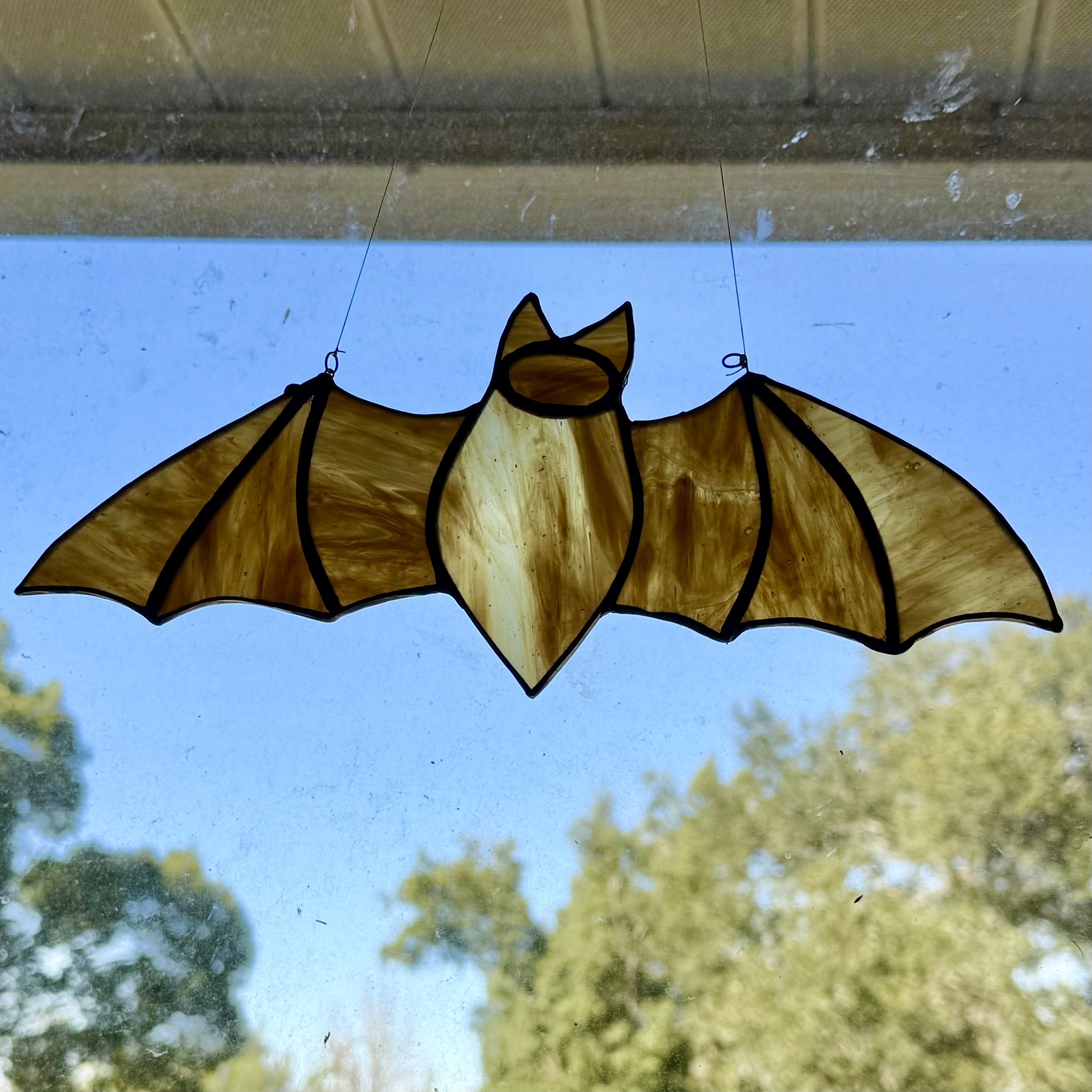 A brown and ivory stained glass bat hanging in a dirty window. Blue skies and green live oak trees are in the soft-focused distance.