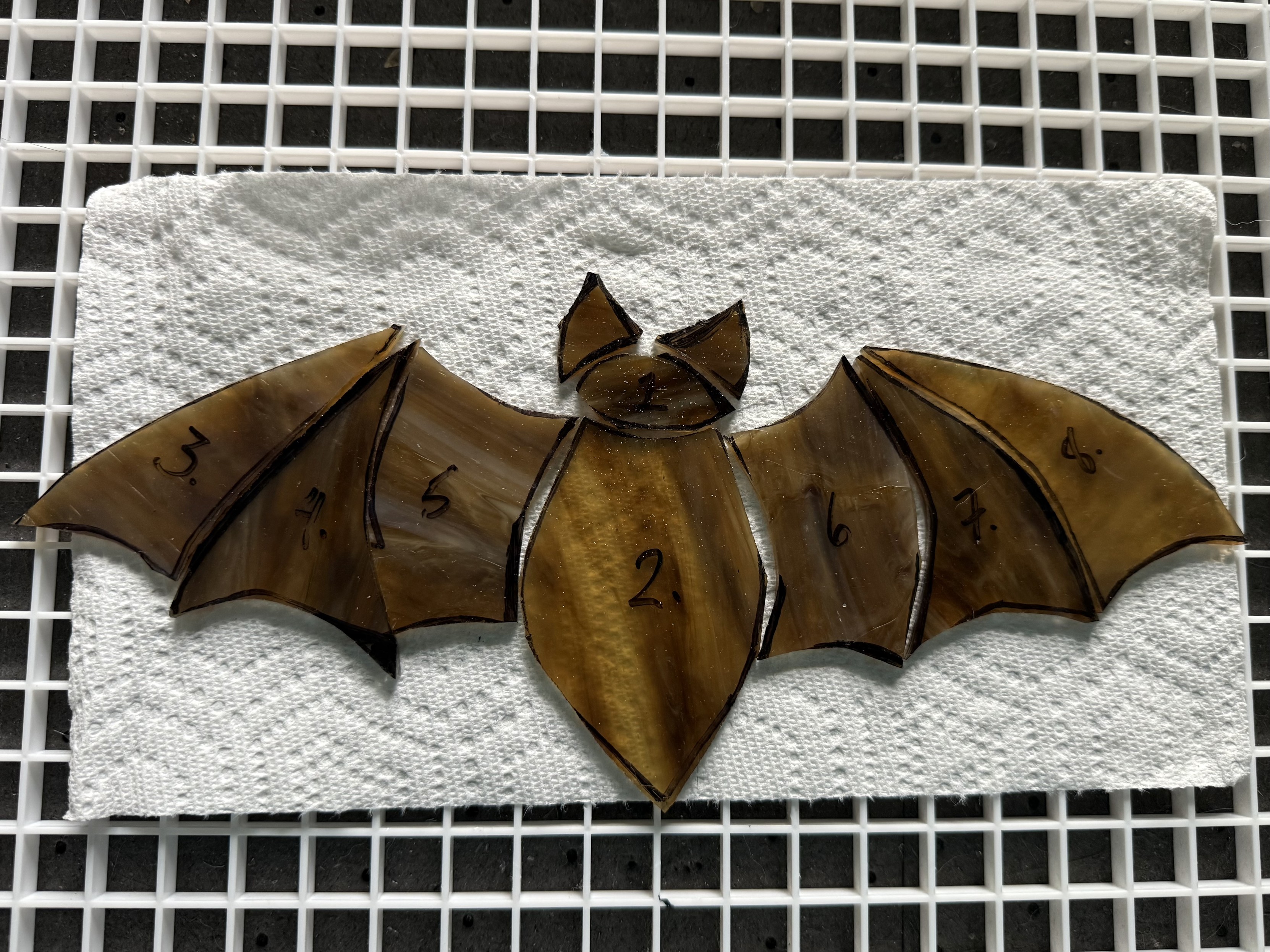 Cut glass pieces arranged as a bat rest on a paper towel half-sheet over a white plastic grid. The bat pieces are numbered for later assembly and have not yet had their edges ground to fine tune the fit.