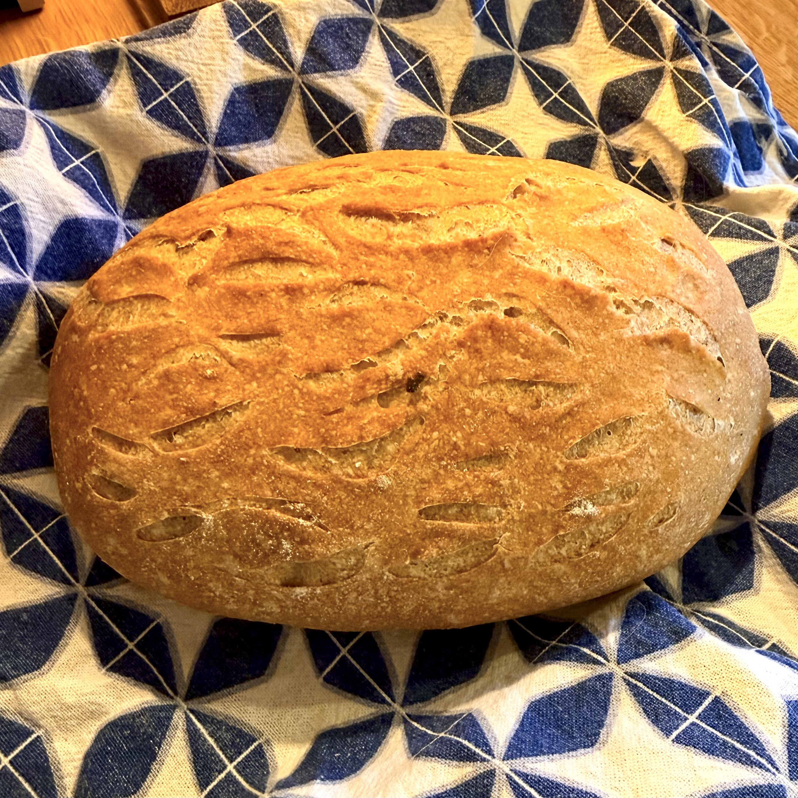 A small oval loaf of golden sourdough bread resting on a tea towel with blue and white geometric pattern.