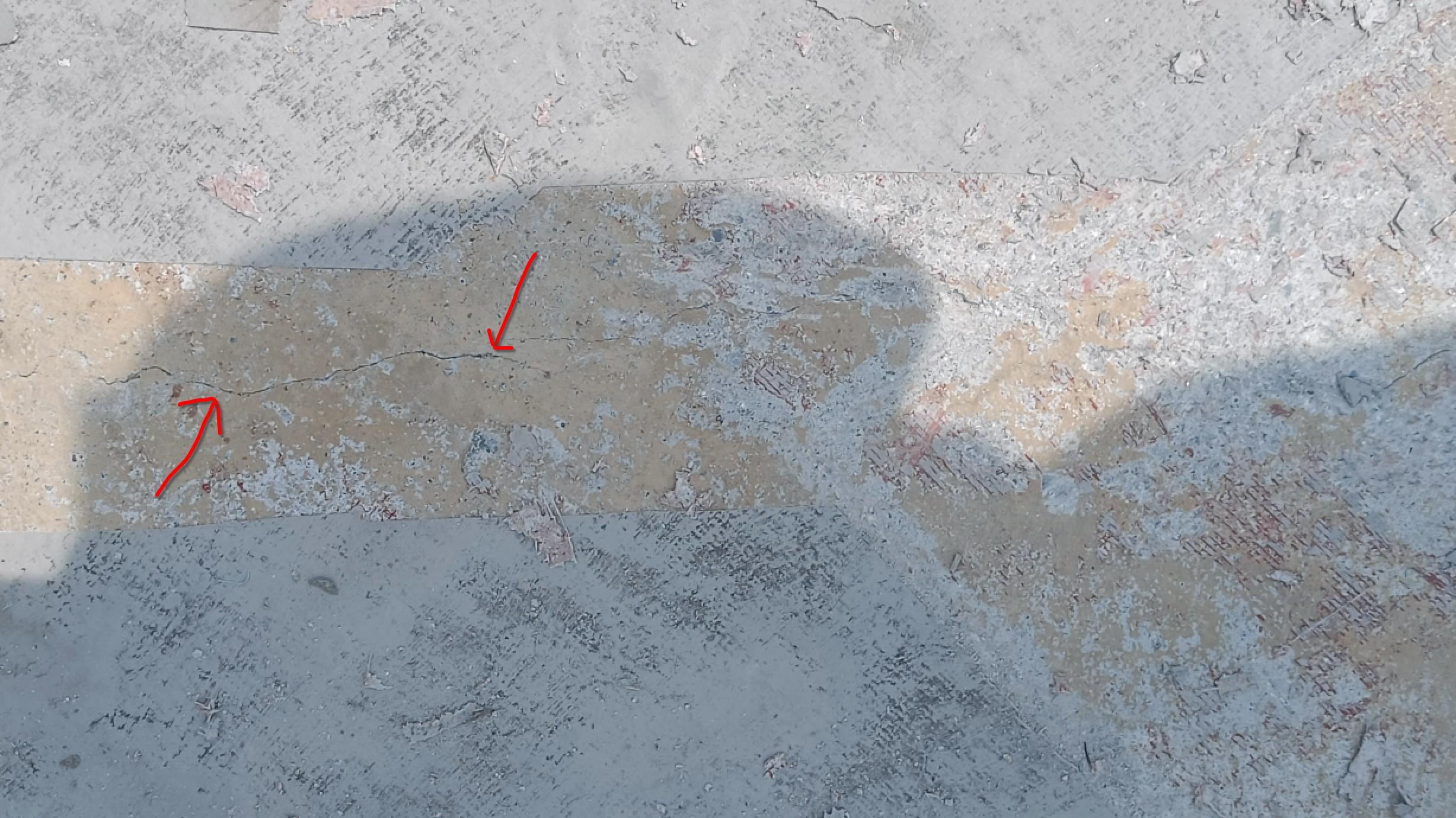 Photo of a beige concrete slab with two red arrows marking a small hairline crack at two points along its length. On either side of the crack are grey edges of the waterproofing (paint) layer applied after curing the slab, here cut away to expose the crack for repair.
