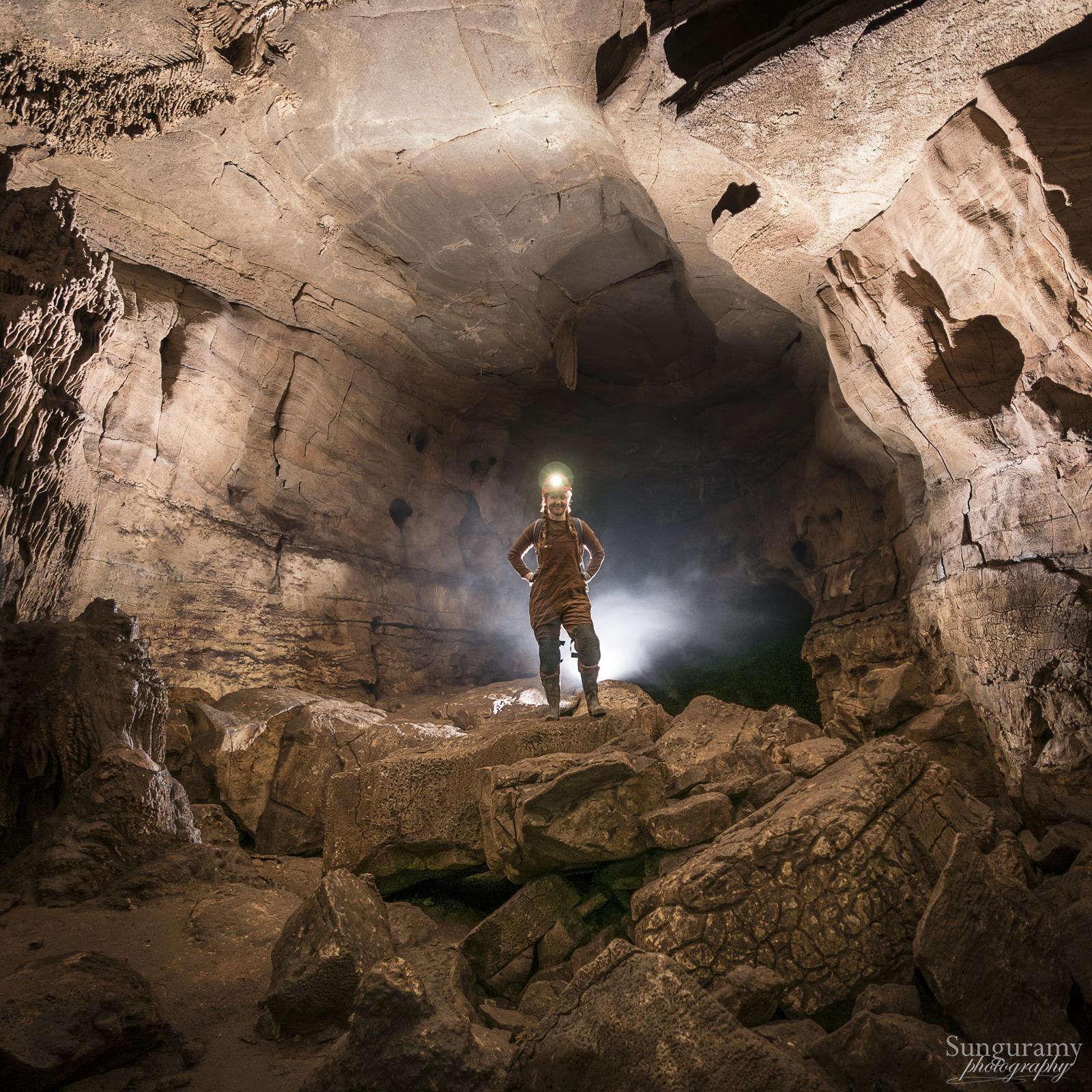Square photograph of a large cave passage. A lone figure in brown coveralls stands in the middle, backlit by a flash. Caves are moist so there is fog / steam in the air coming which highlights the figure and although the cave is brown as well, helps them stand out. Lots of big boulders on the floor, and a hint of formations on the right side.