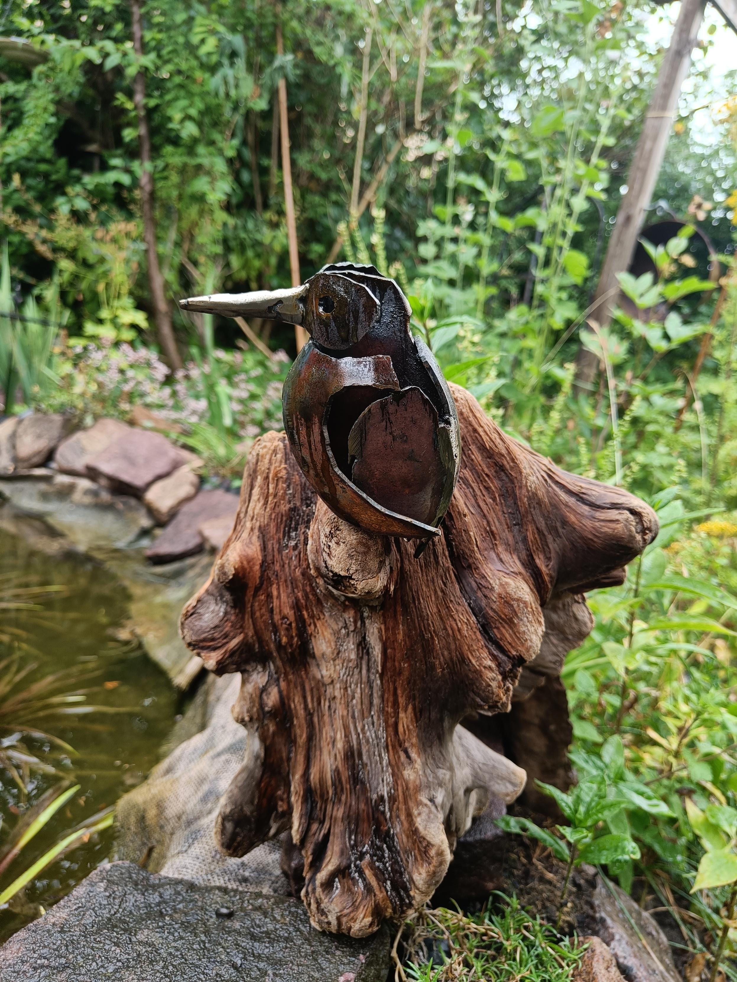A Kingfisher made of weathering steel sits perched on a driftwood root.