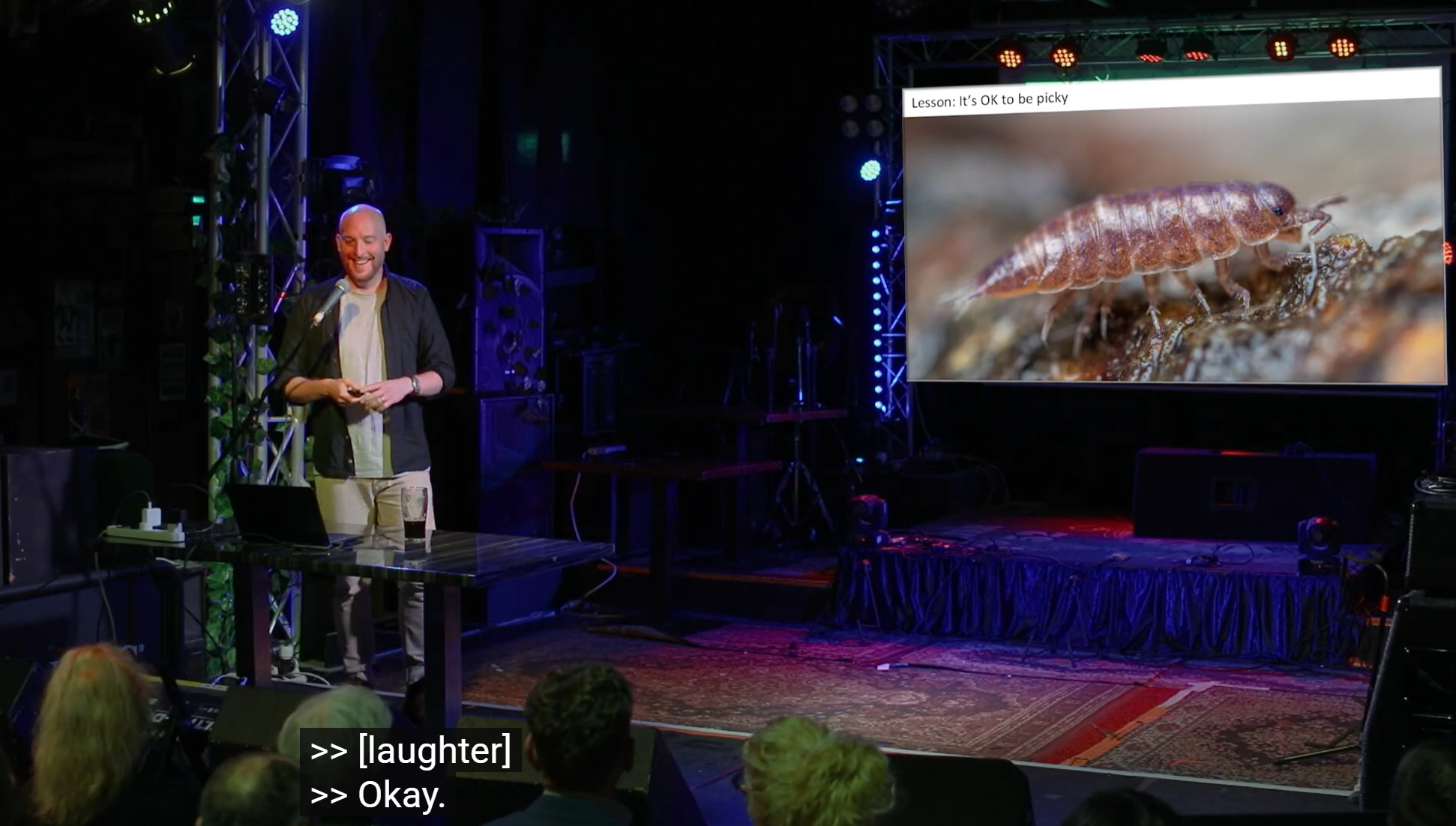 A photo of me standing on a stage, giving a talk about soil animals. On the screen behind me is a photograph of an iridescent purple woodlouse.
